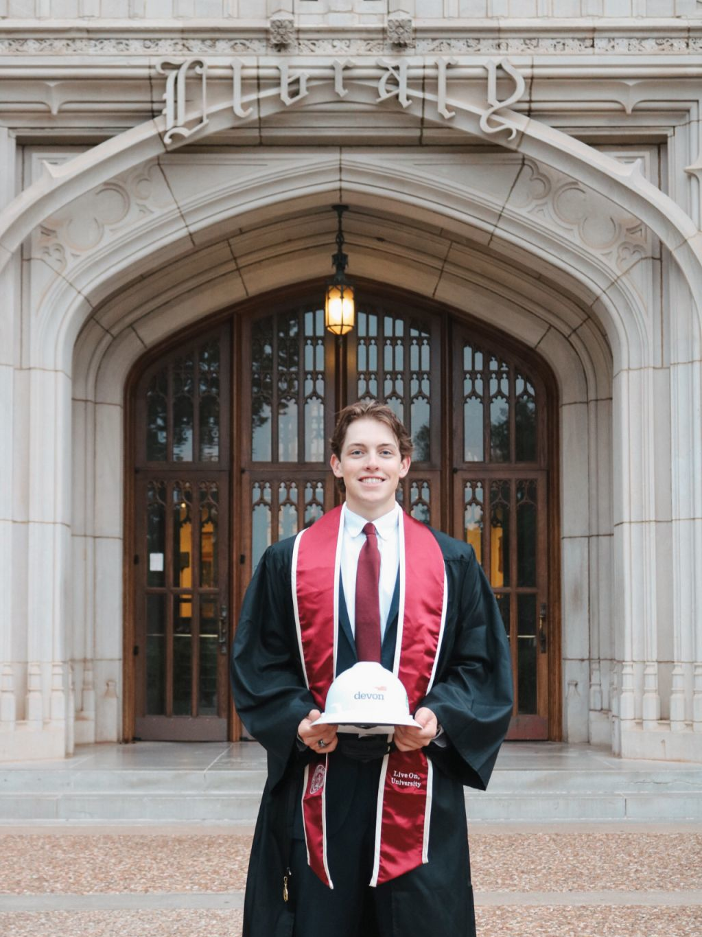 Lucas Livingstone graduates OU in his regalia holding a Devon hardhat.